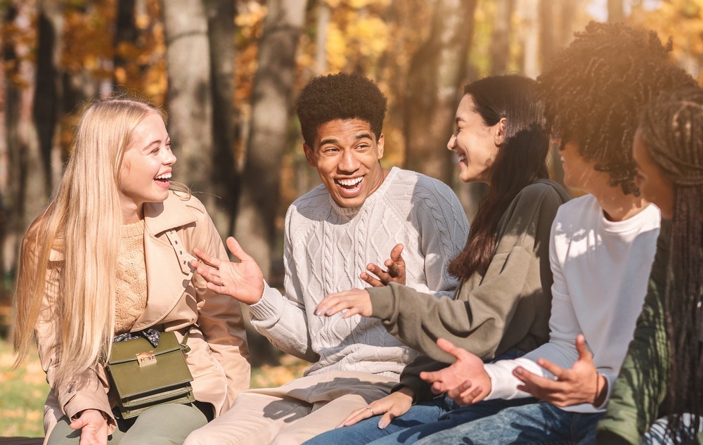 A group of young adults sitting on a bench outdoors, laughing and talking together in a park with autumn trees in the background.